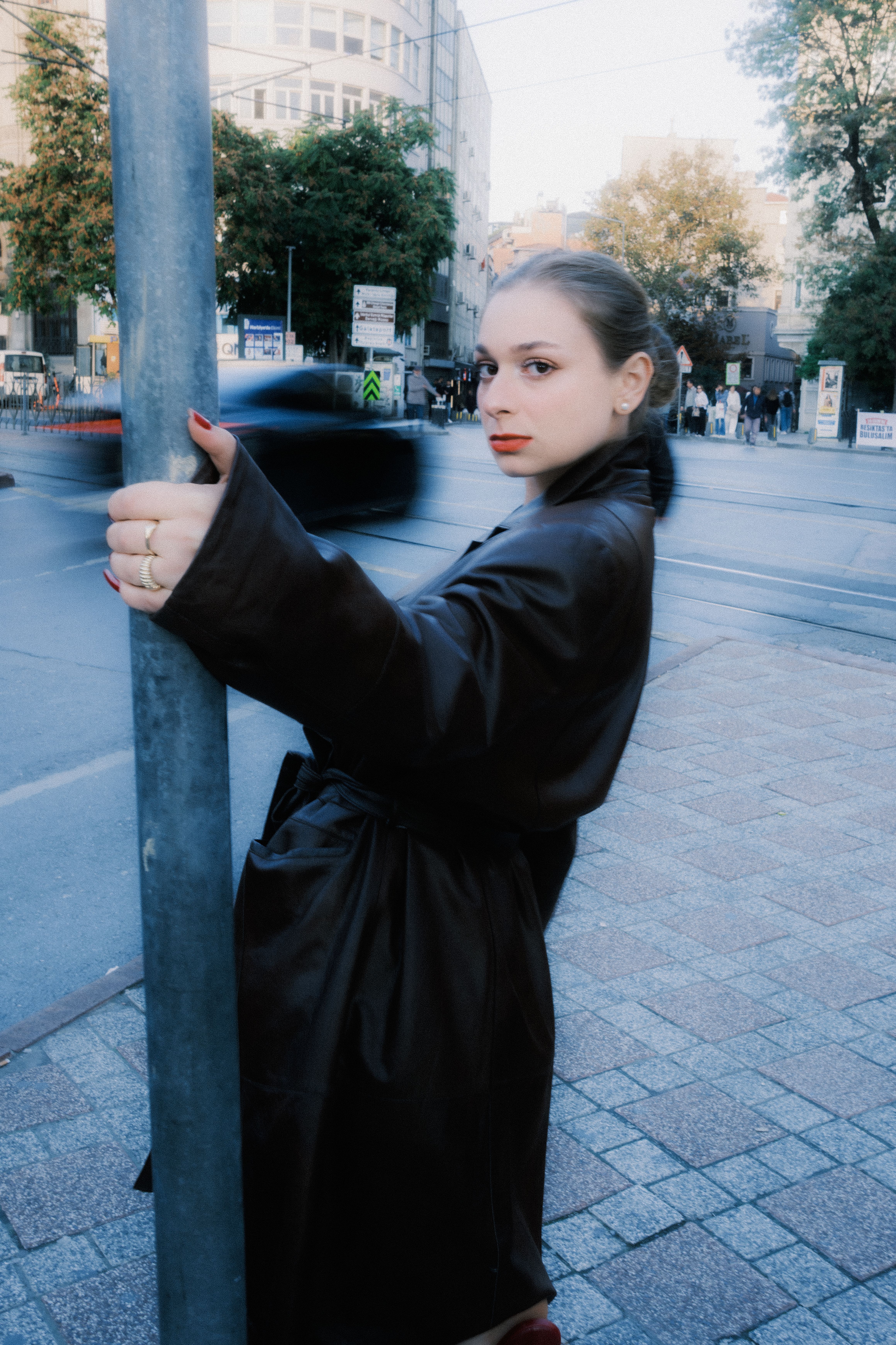 A fashionable woman in a dark trench coat holds a pole on a bustling city street. Urban lifestyle vibe.