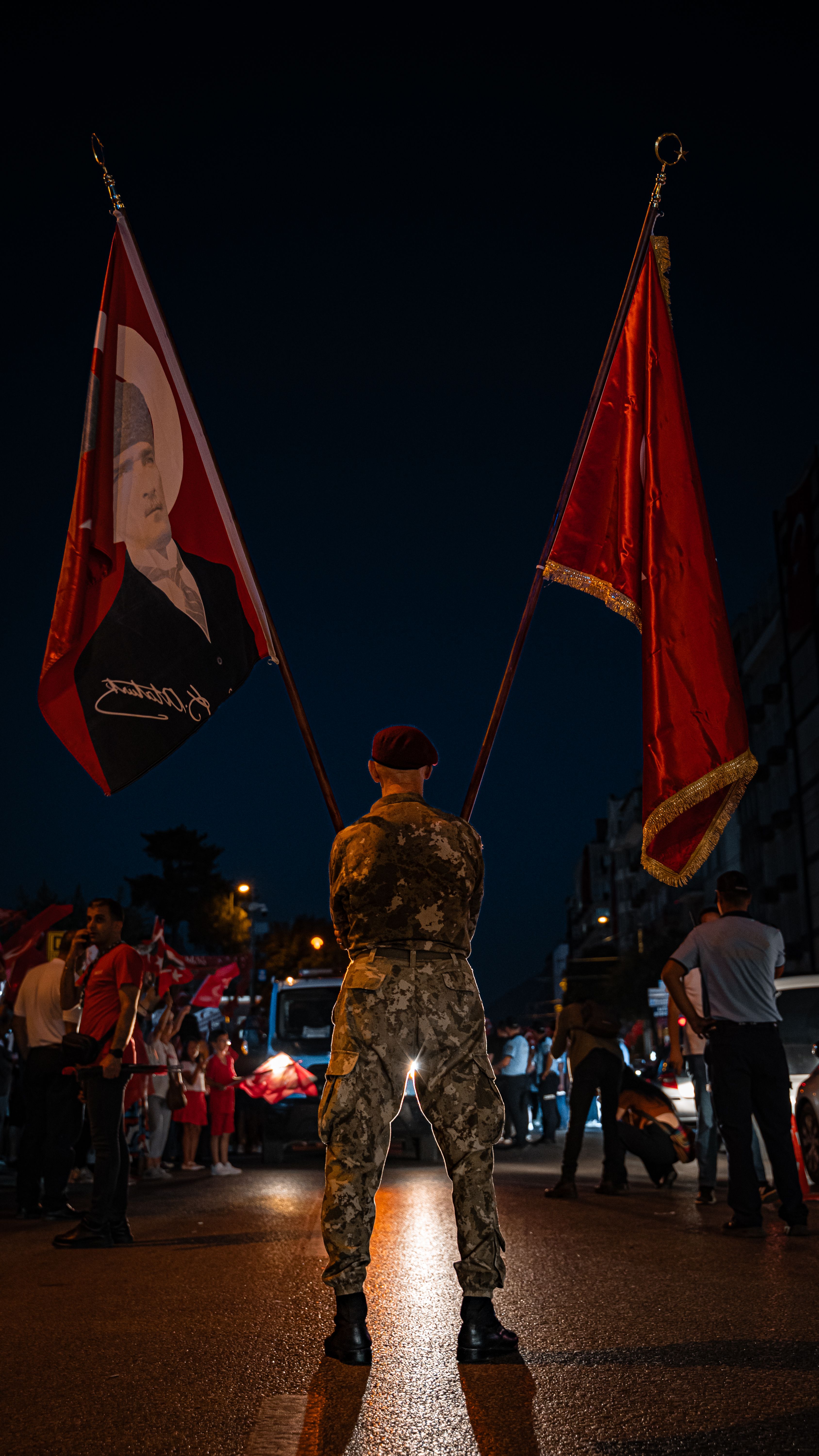 Soldier holding flags during a night event with a city backdrop, showcasing national pride.