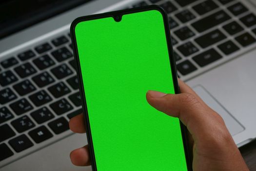 Close-up of a smartphone with a green screen held by a hand over a laptop keyboard.