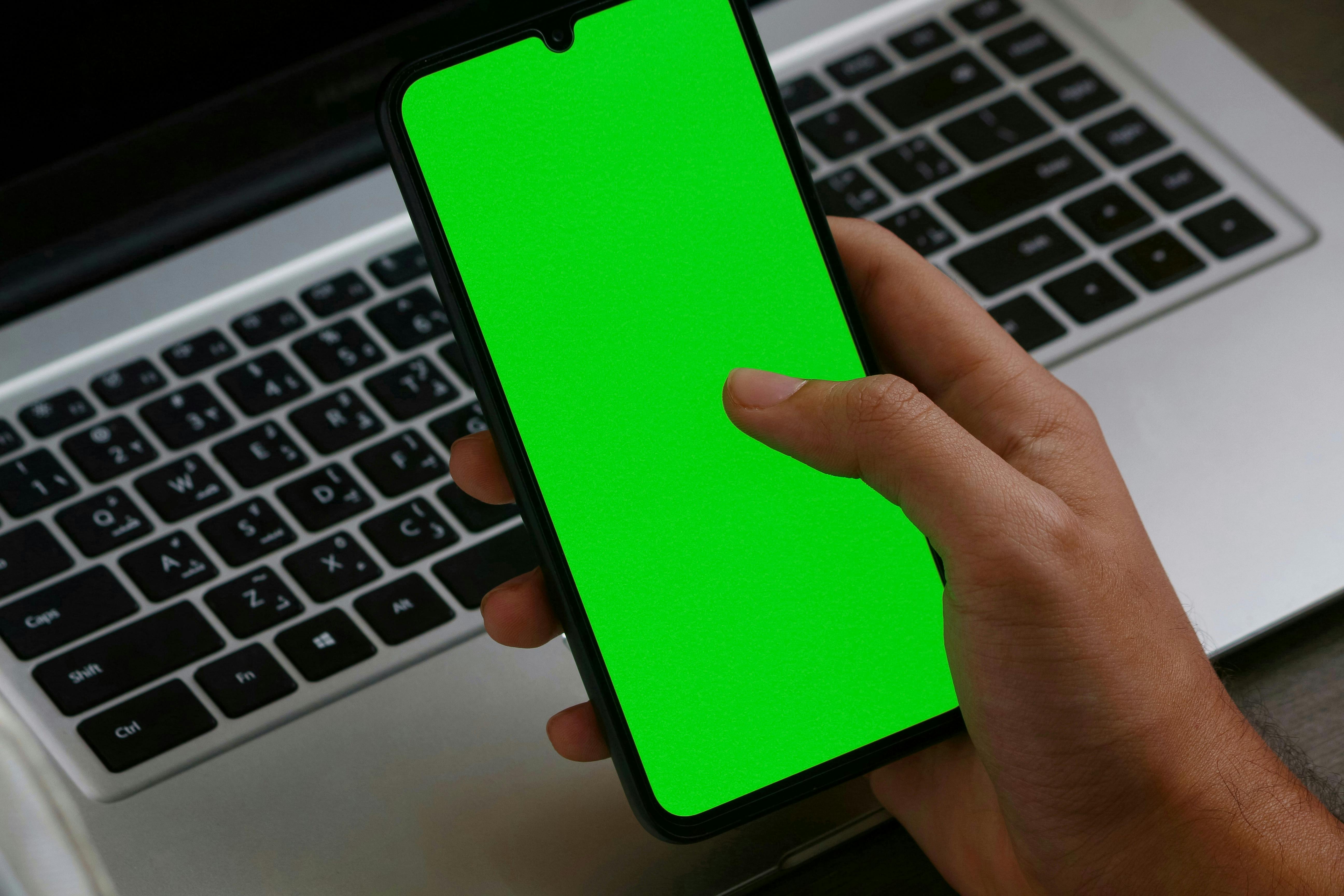 Person Holding a Smartphone with Green Screen Near a Laptop