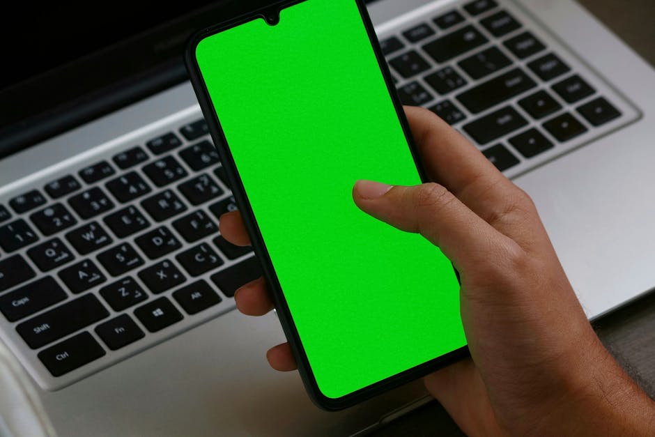 Person Holding a Smartphone with Green Screen Near a Laptop