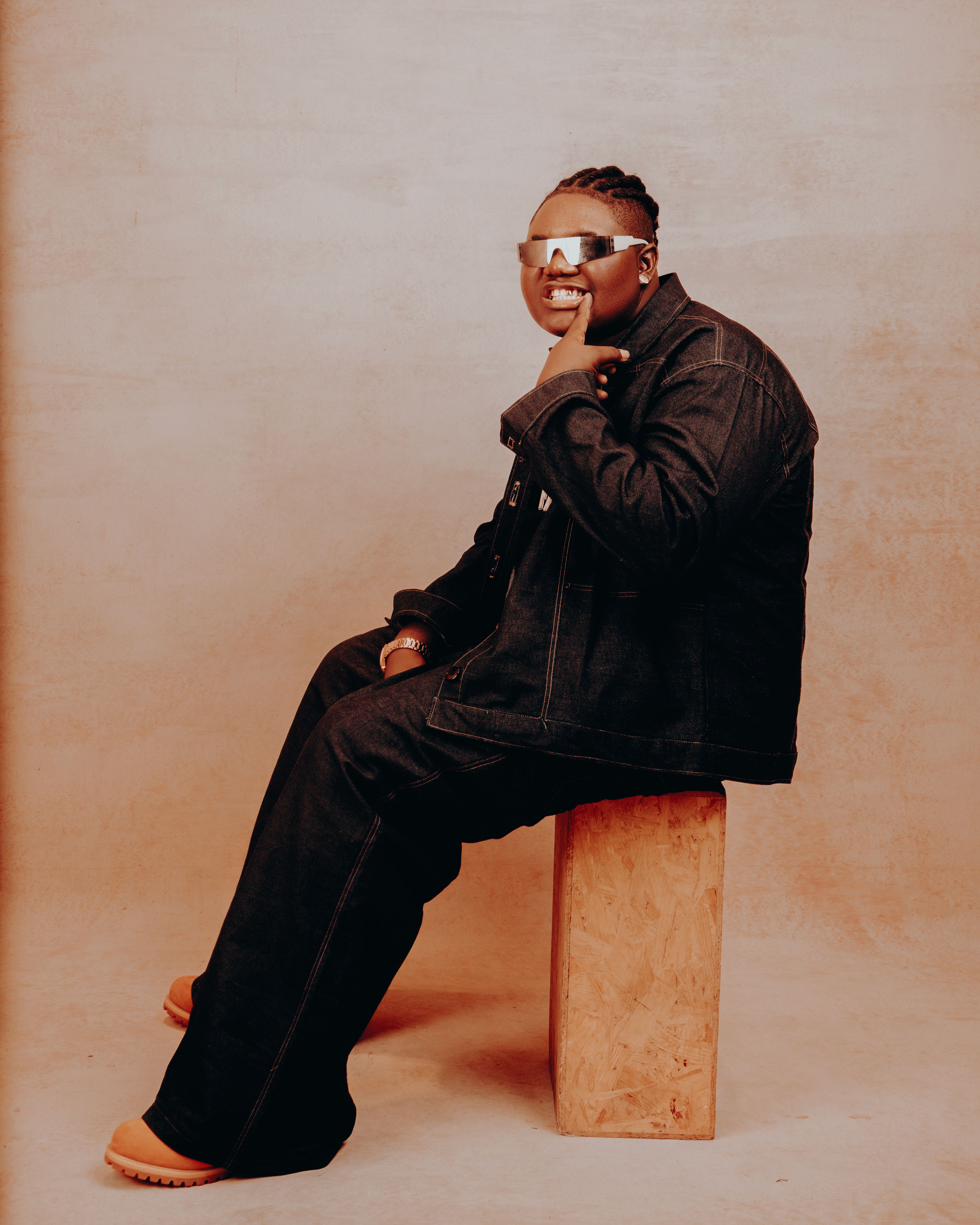 A fashionable adult seated on a stool wearing dark denim and reflective sunglasses.