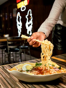 Close-up of flavorful ramen being lifted with chopsticks in a cozy restaurant with neon lights.