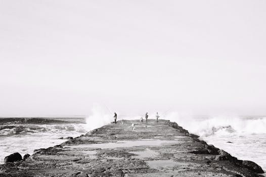 Black and white photo of people fishing on a rocky pier with waves crashing.