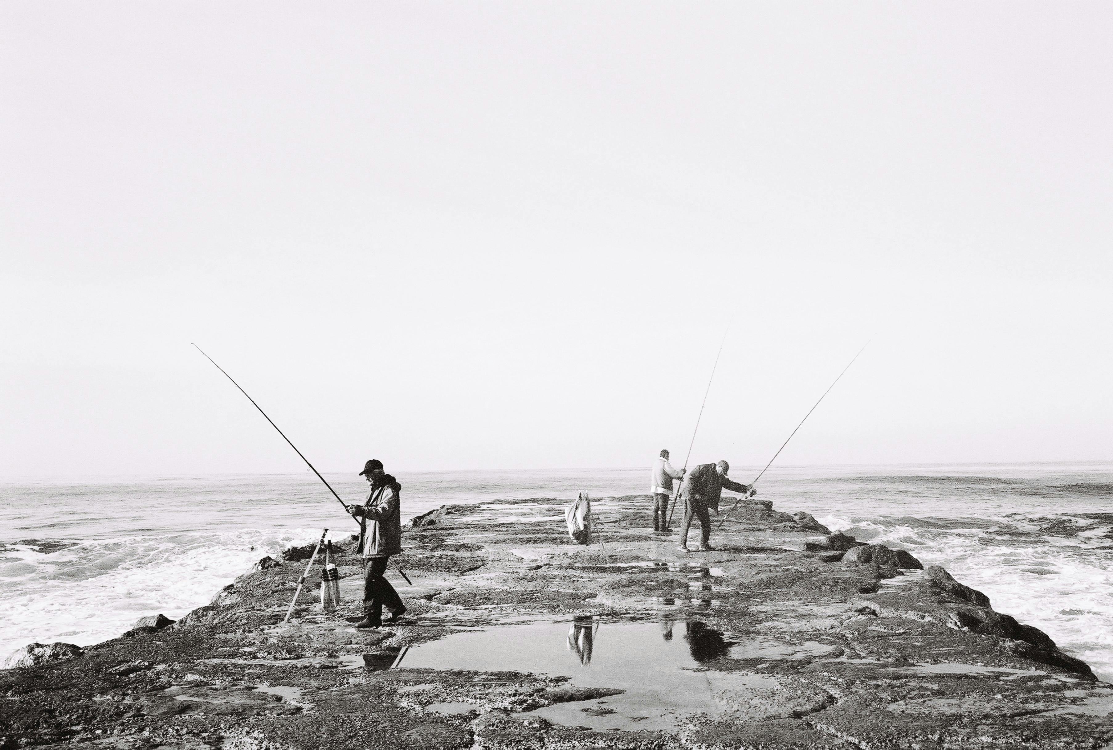 Three men fishing on a rocky pier by the sea in black and white.