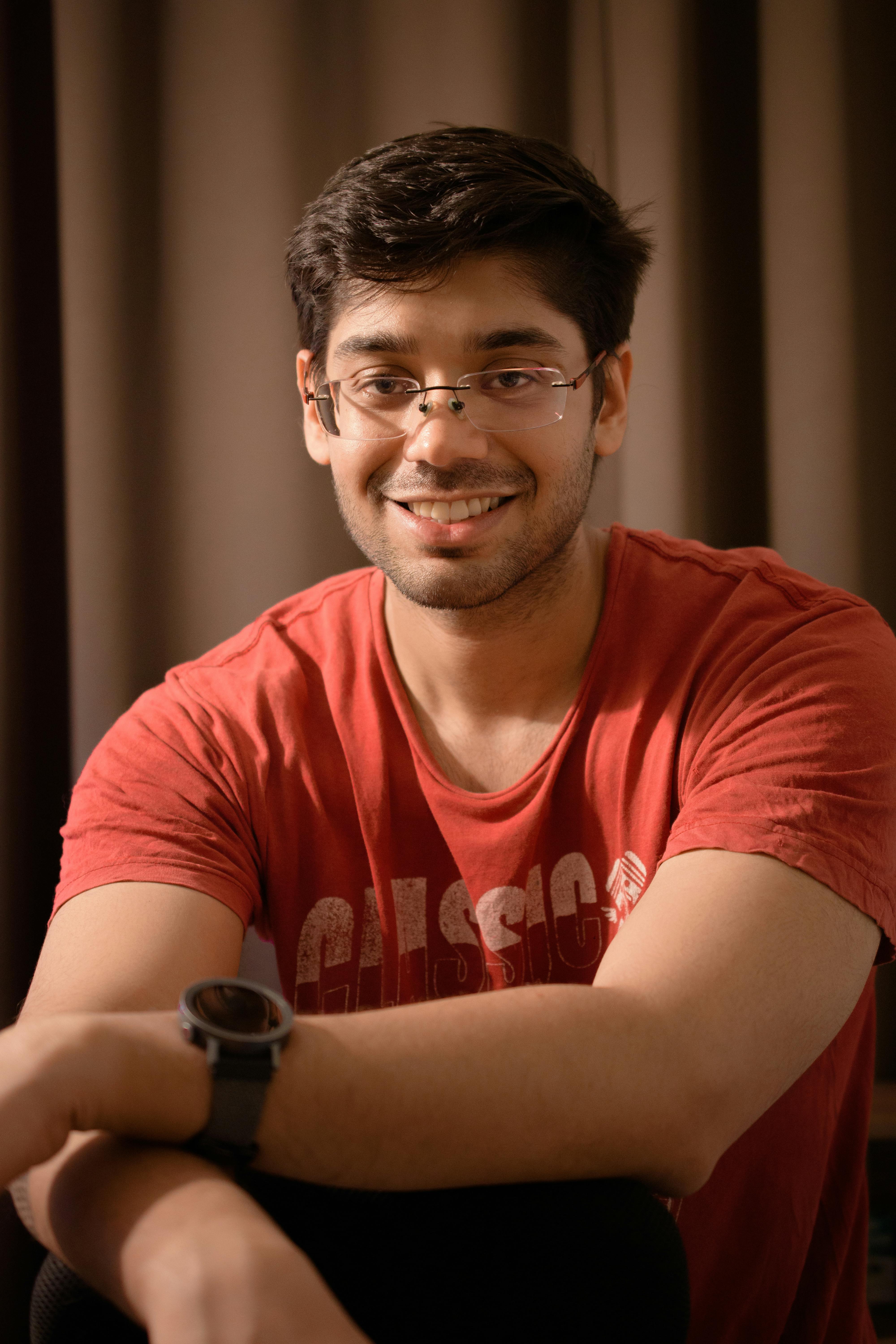 A young man wearing glasses and a red t-shirt smiles warmly, sitting indoors.