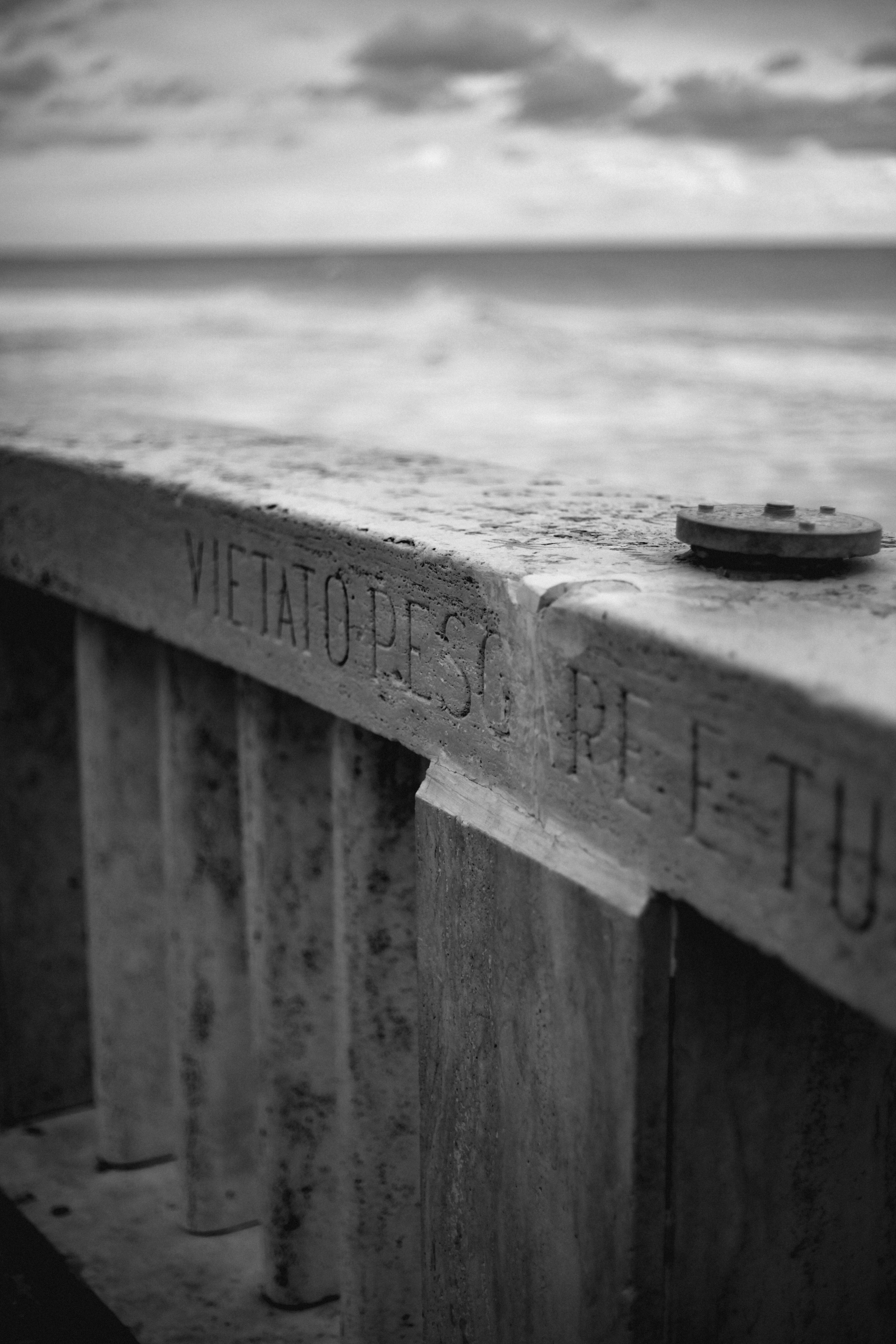 Moody black and white photo of a coastal barrier with inscription 'Vietato Pescare.'