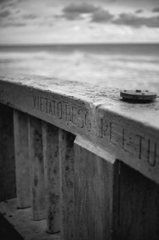 Moody black and white photo of a coastal barrier with inscription 'Vietato Pescare.'
