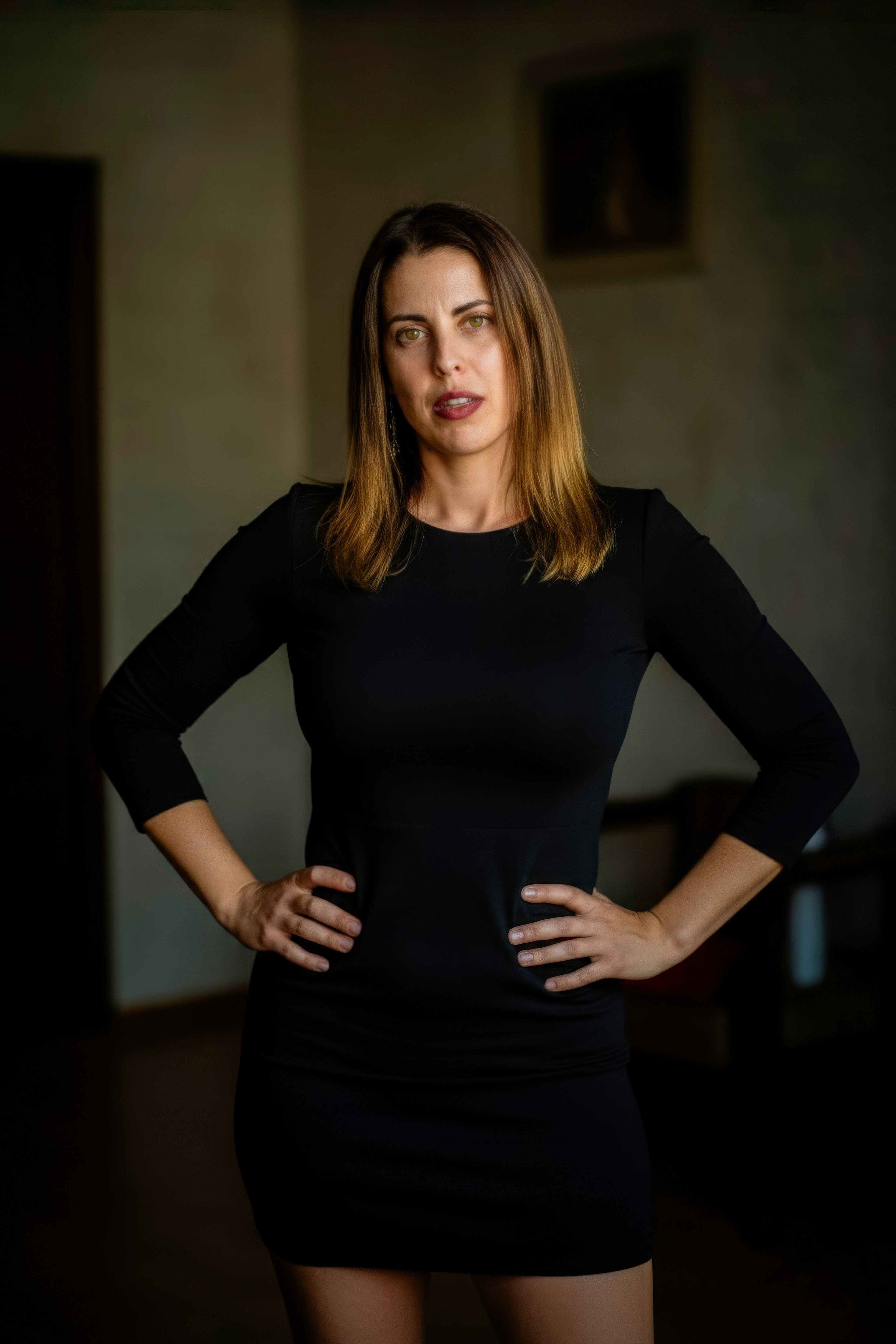 A confident woman wearing a little black dress stands indoors with hands on her hips.