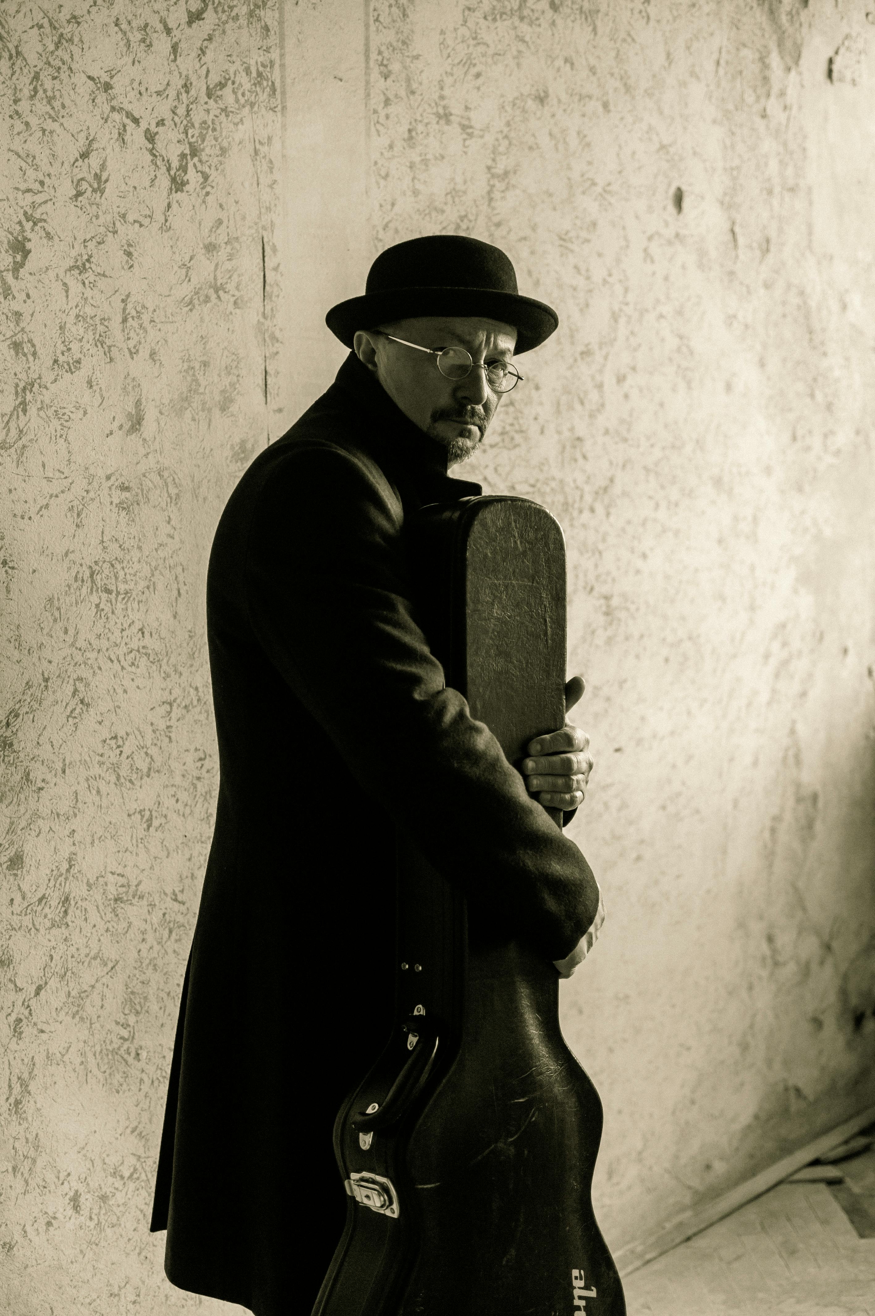 A man in a fedora holds a guitar case in a vintage-styled portrait.