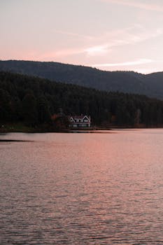 Tranquil view of a lakeside house surrounded by autumn forest during sunset.