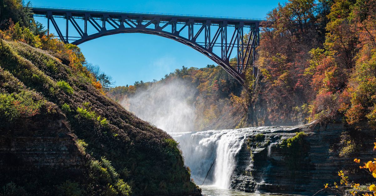 A stunning autumn landscape featuring a waterfall under a historic bridge.