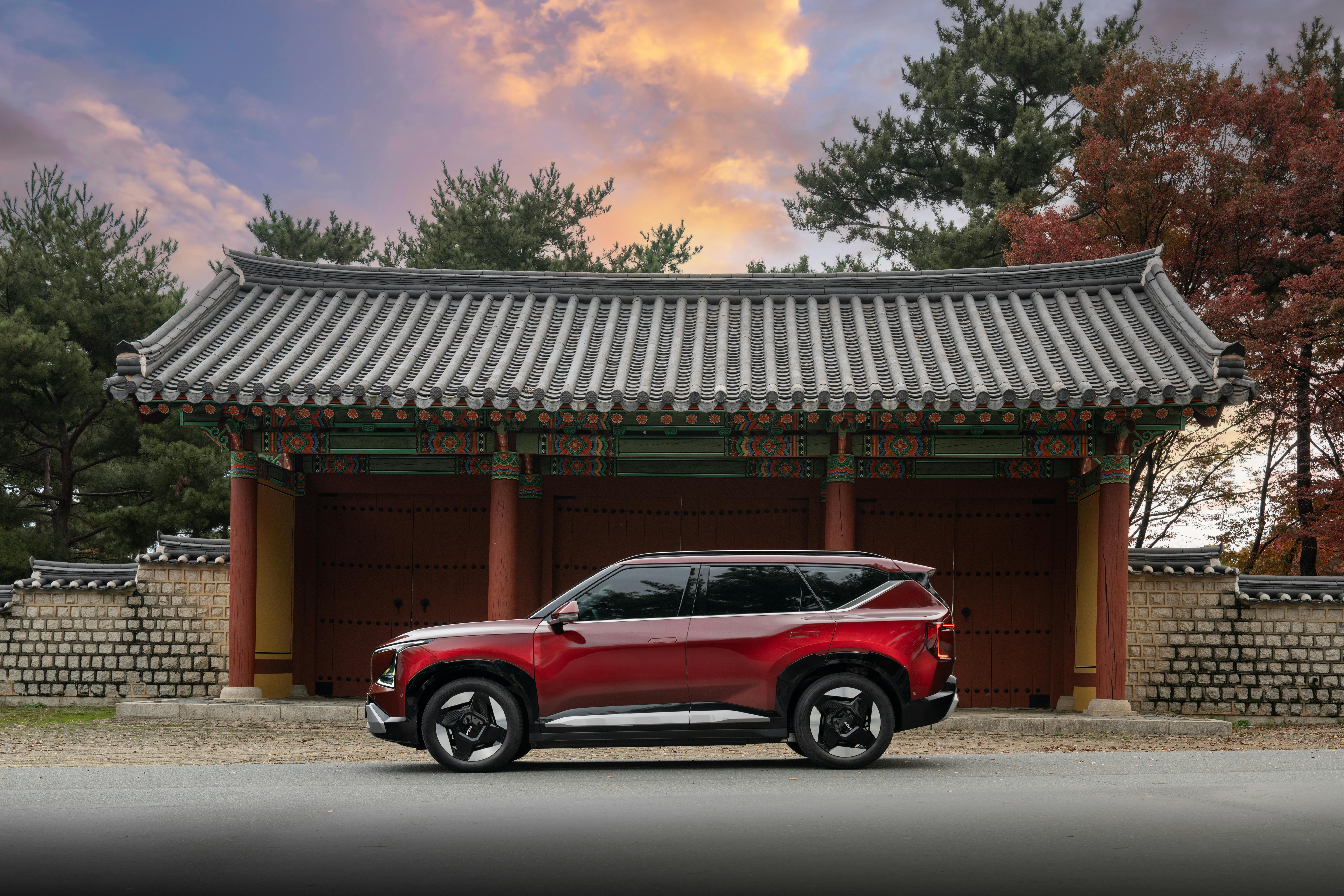 A sleek red SUV is parked in front of a traditional Korean gate with autumn foliage and a vibrant sunset.