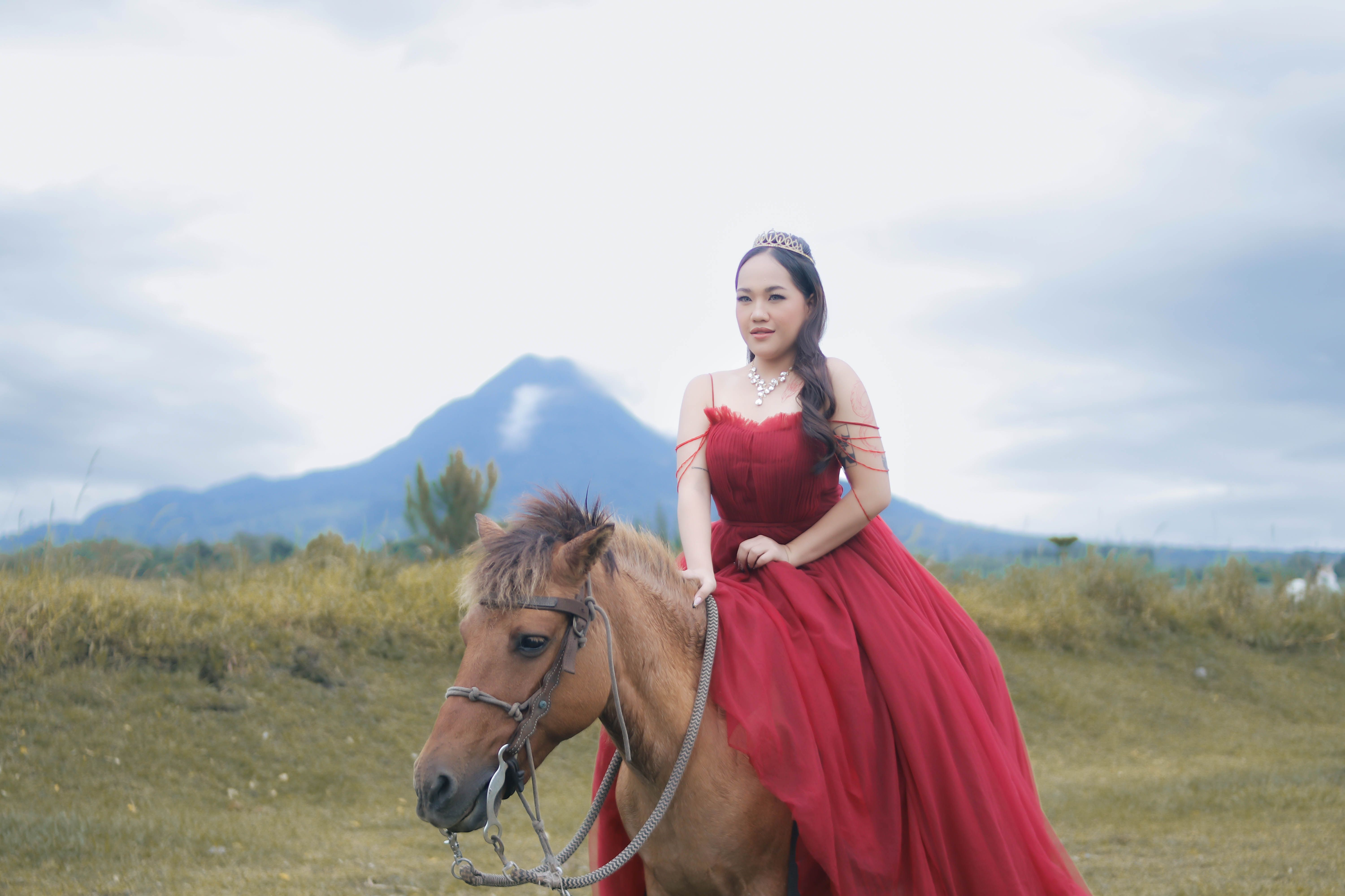 A woman in a red gown and tiara rides a horse with a majestic mountain backdrop.