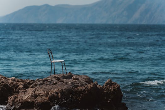 A solitary chair on a rocky shore overlooking a vast ocean with mountains in the distance.