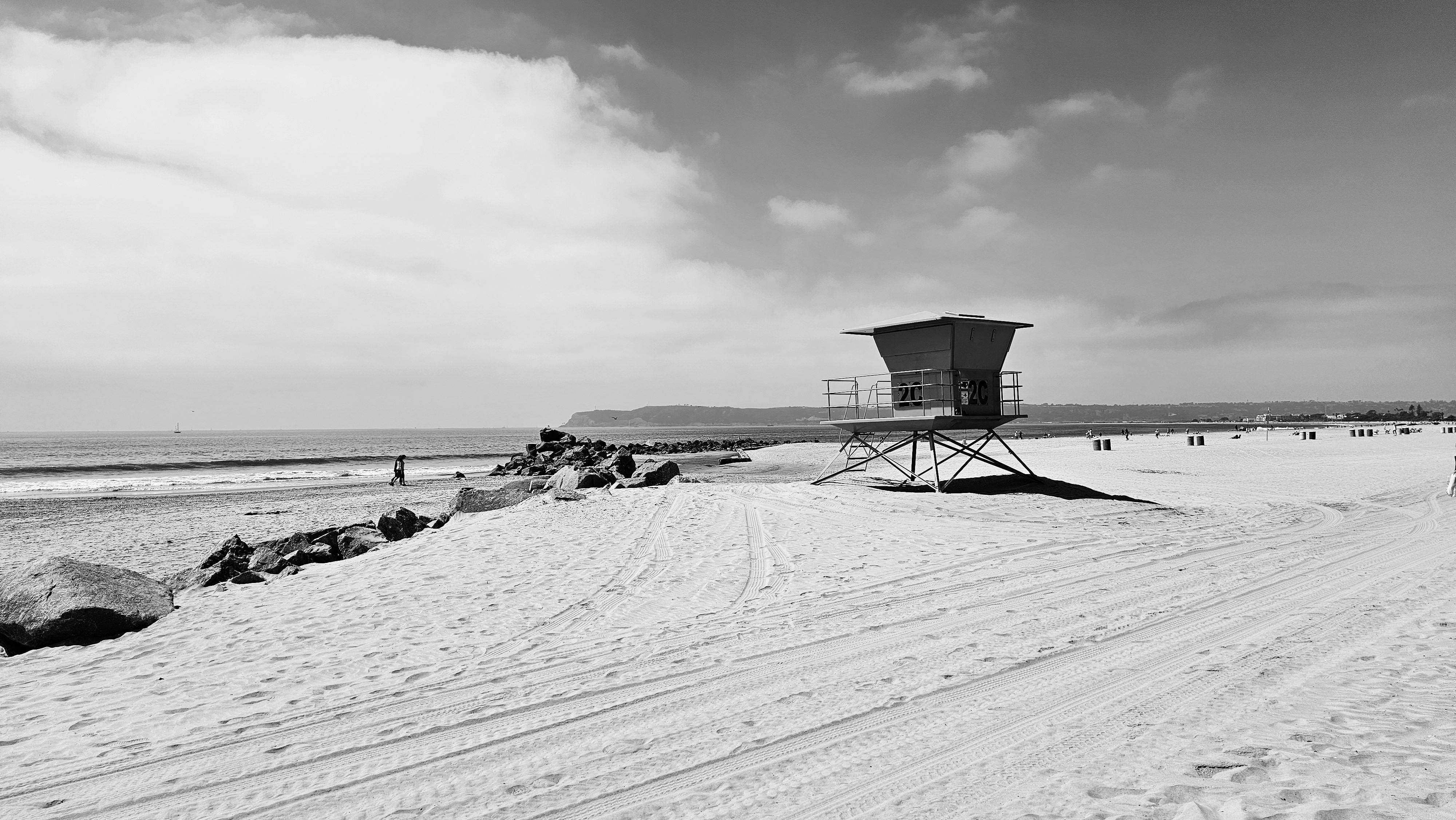 A serene black and white image of a beach with a lifeguard tower and distant walkers.
