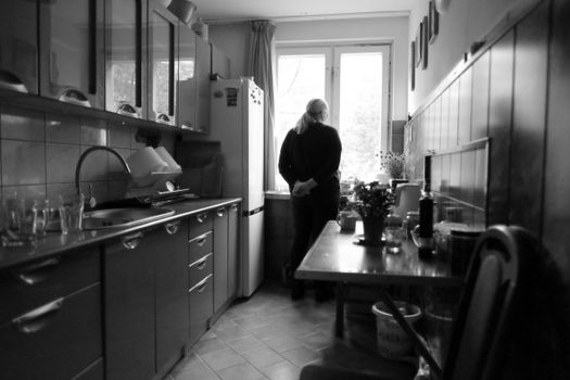 Black and white image of a person in a Polish kitchen, gazing out the window, creating a contemplative mood.