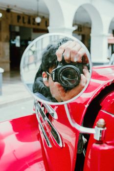 Photographer capturing reflection in vintage car mirror with modern camera.
