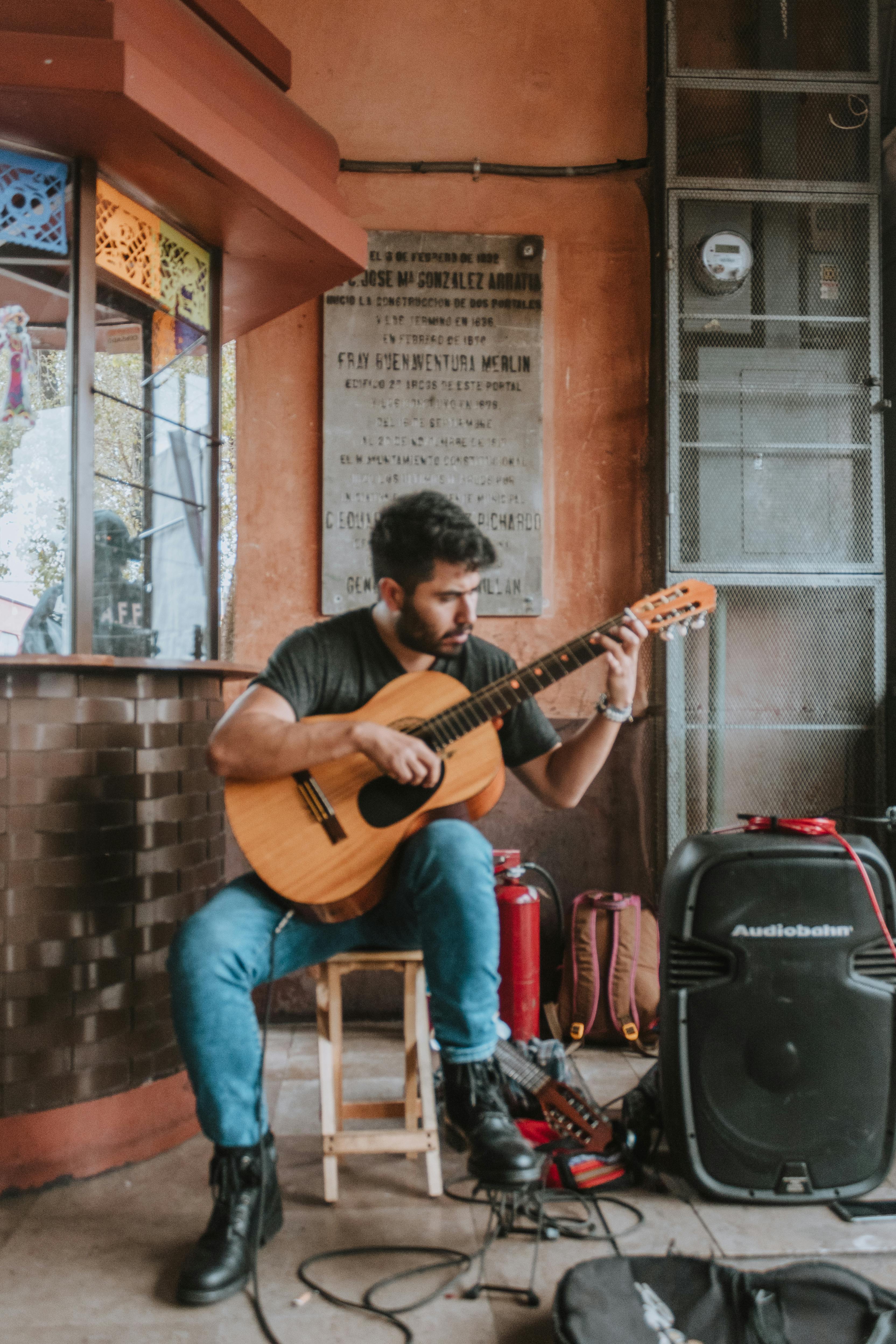 Musician playing guitar on a street, surrounded by equipment and casual setting.