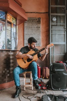 Musician playing guitar on a street, surrounded by equipment and casual setting.