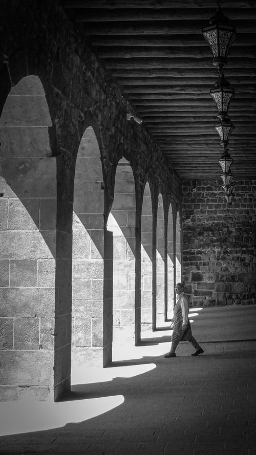 Free A person walks through a sunlit stone corridor with arches casting dramatic shadows. Stock Photo