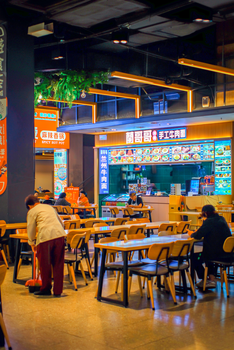 Colorful Asian food court with diners enjoying meals and staff cleaning. Vibrant, modern indoor scene.
