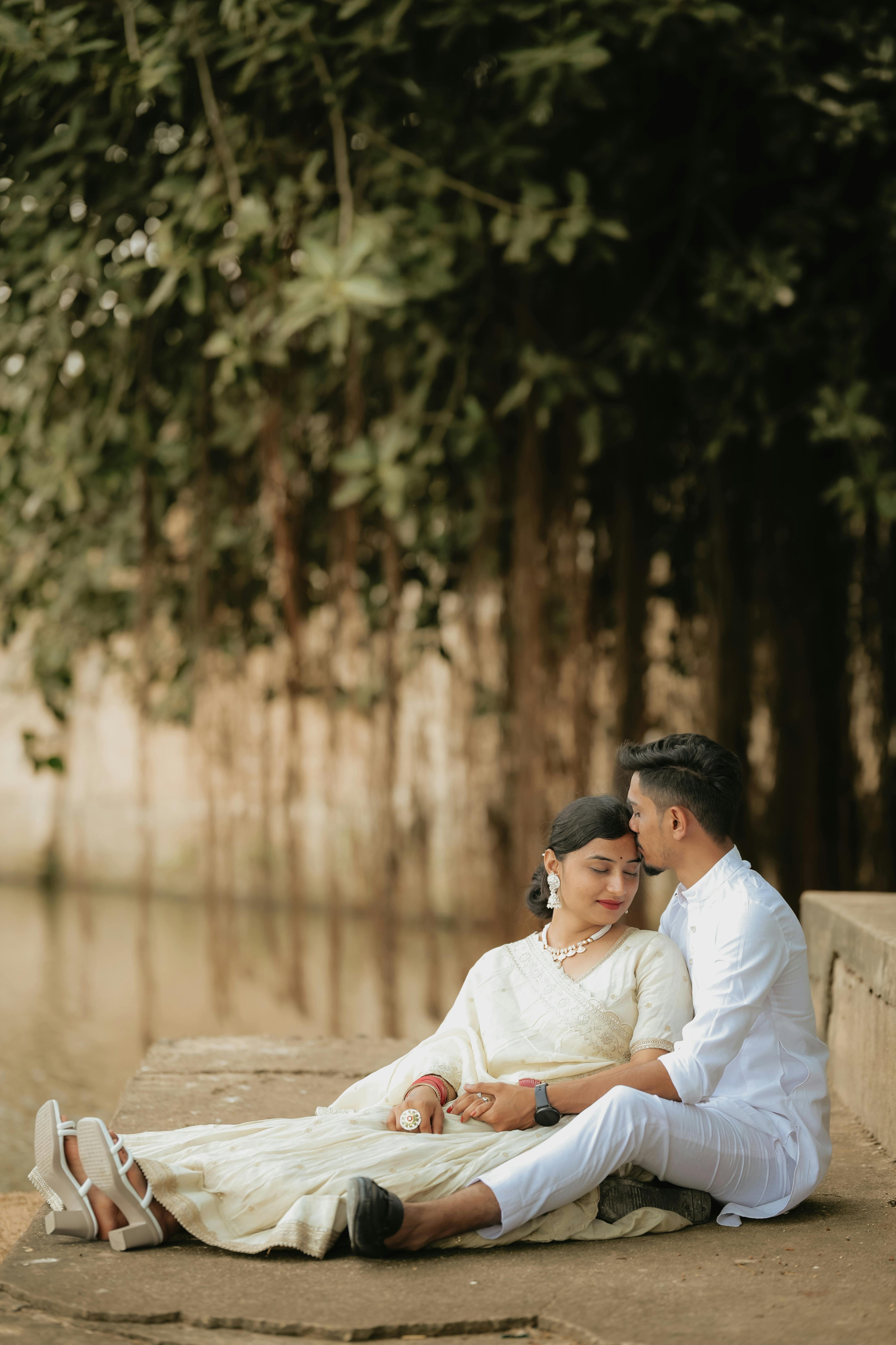A couple in white attire sit closely under a banyan tree, reflecting romance.
