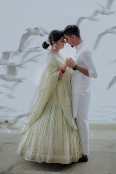 A romantic couple in traditional attire shares an intimate moment by the sea amidst flying seagulls.