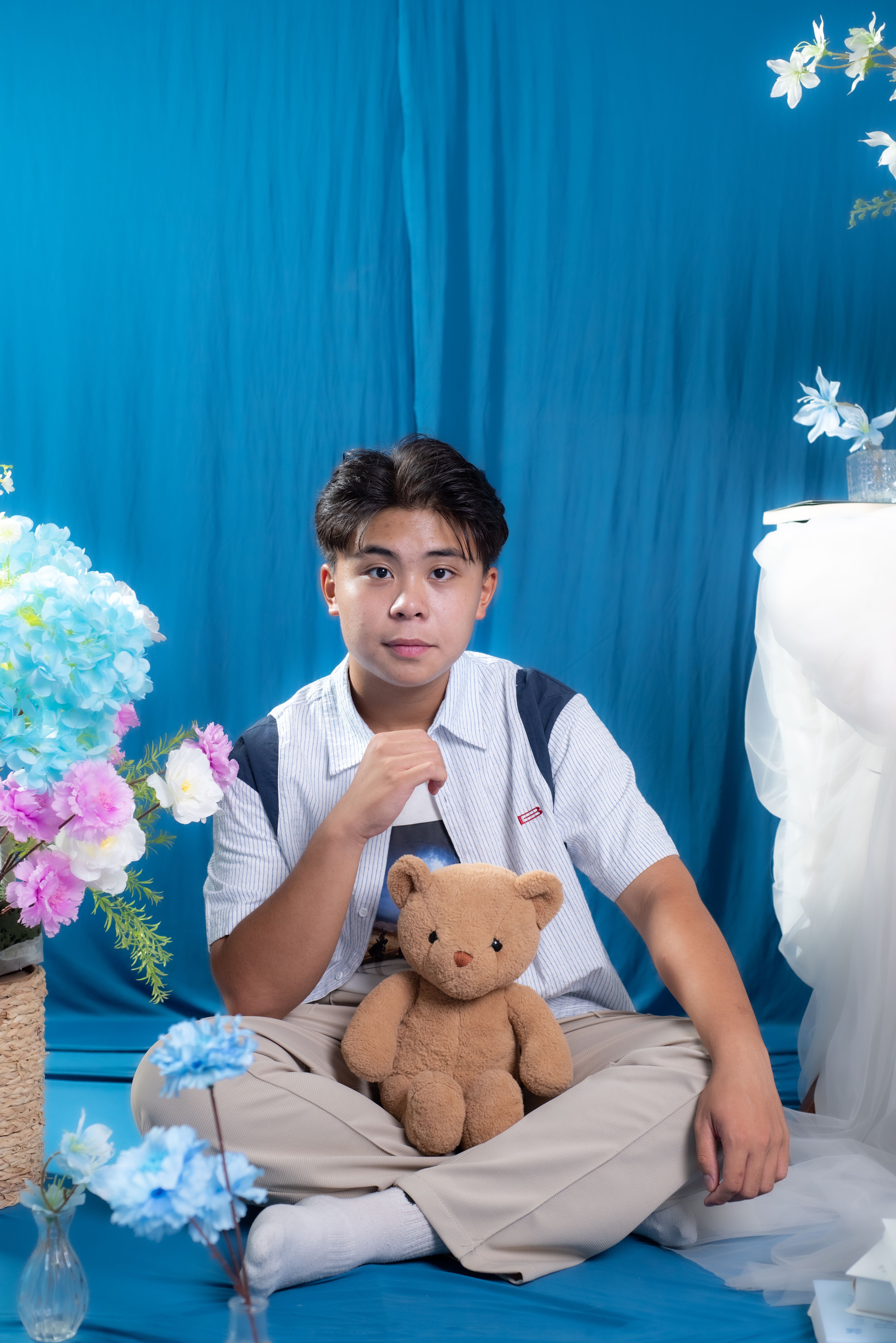 Young man posing with a teddy bear amidst vibrant floral decorations against a blue backdrop.