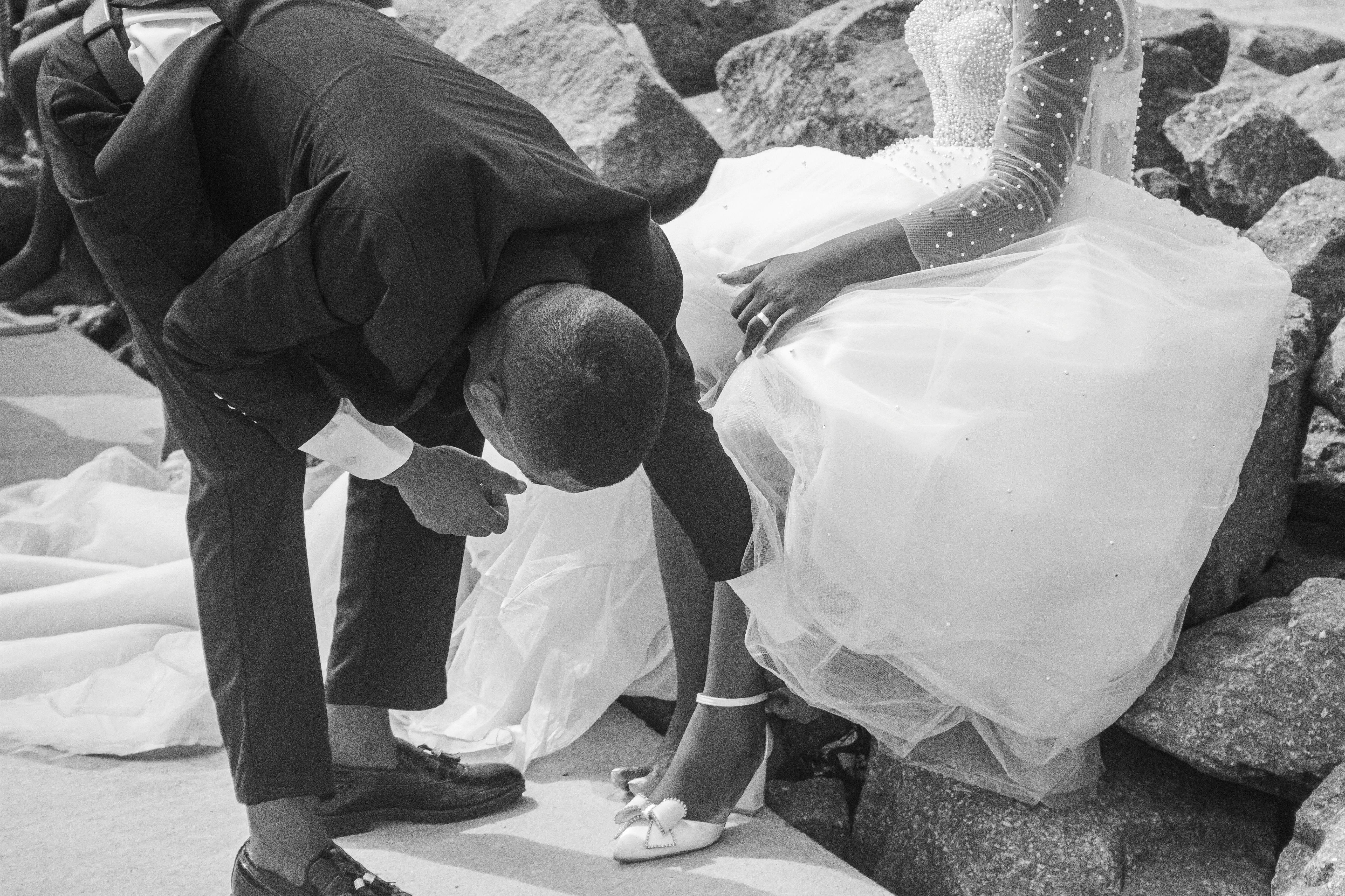 A groom helps adjust the bride's shoe during a stylish outdoor wedding by the rocks.