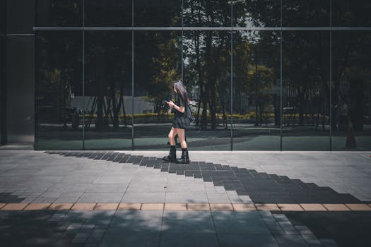 A young woman checks her phone while walking past a glass building. Bright sunny day reflects urban lifestyle.