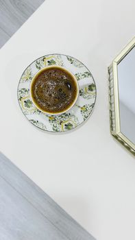 Top-down view of coffee in a floral saucer near a gold-edged mirror on a white table.