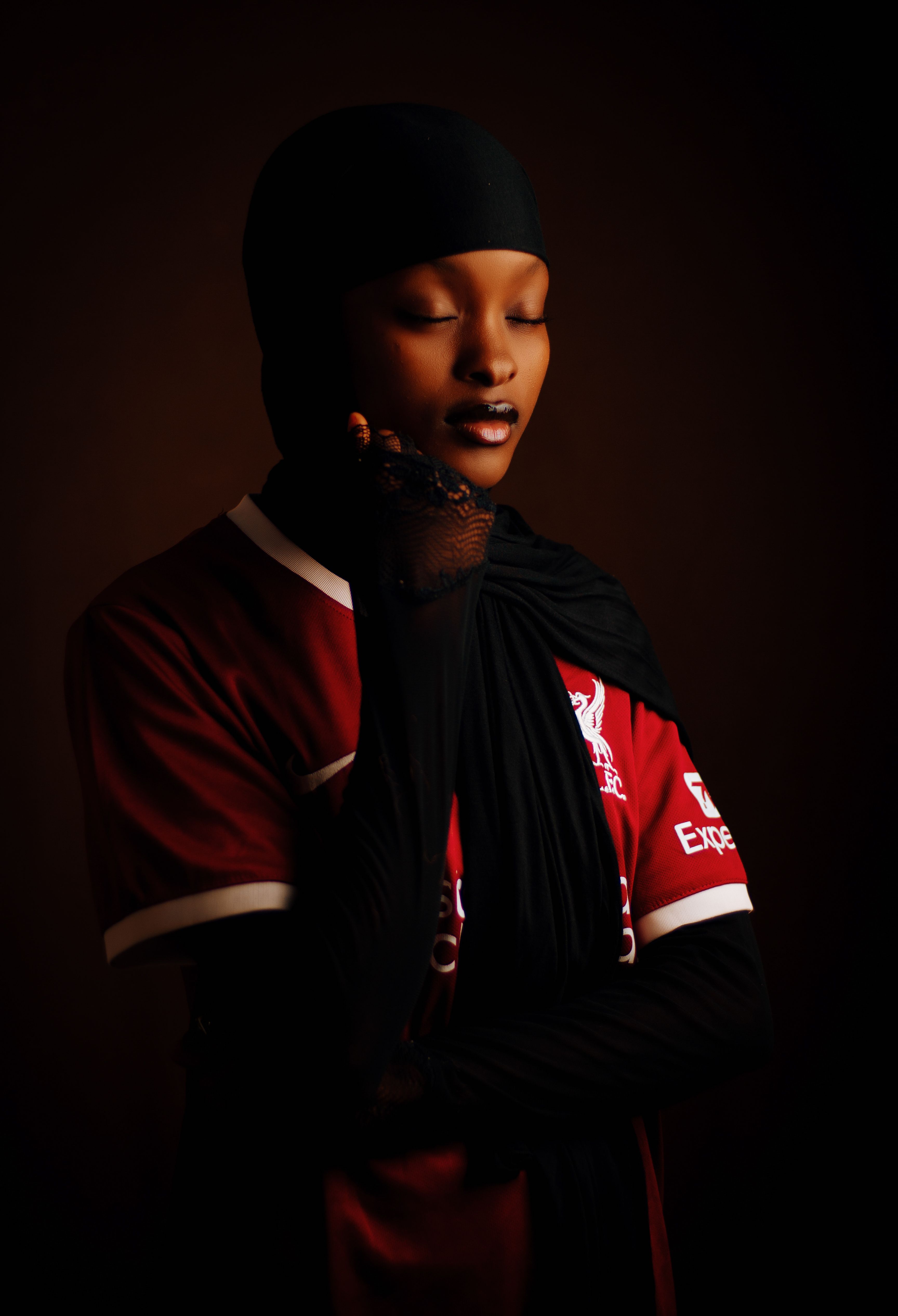 Female in hijab with sports jersey and serene expression against dark background.