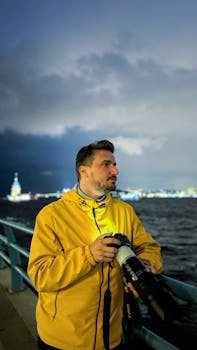 A person with a camera by the waterfront during dusk, city lights in background.