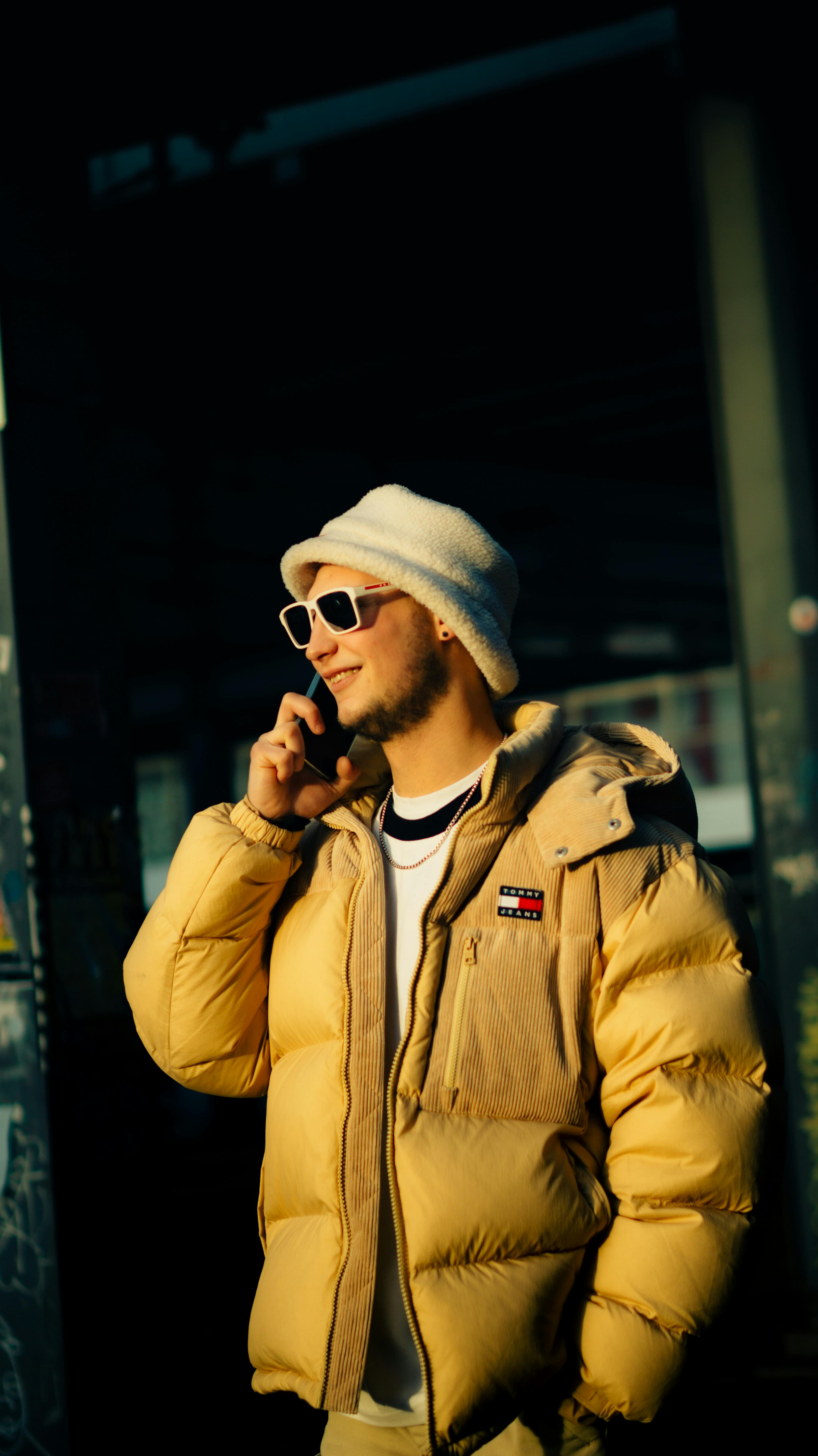 A young man in a beige jacket and sunglasses talking on his phone on a sunny day.