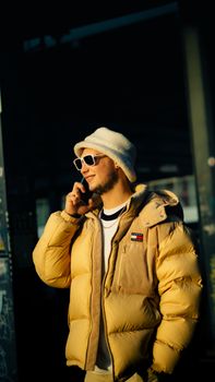 A young man in a beige jacket and sunglasses talking on his phone on a sunny day.