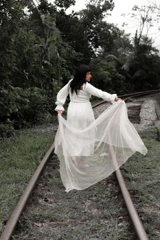 A woman in a flowing white dress walks along railroad tracks surrounded by greenery.