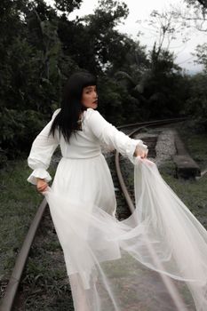 A woman in flowing white dress walks enchantingly along a railway in a lush forest setting.