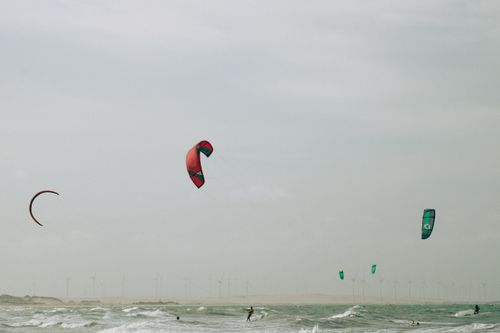 Free Kitesurfers enjoy thrilling rides on the ocean waves with wind turbines in the distance, a blend of adventure and renewable energy. Stock Photo