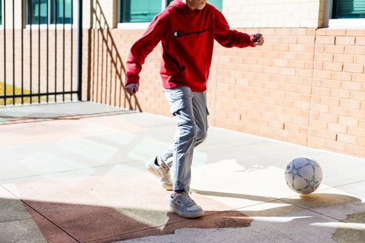 A teenager in a red hoodie playing soccer on a sunny day in a courtyard.