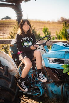 Smiling girl sitting on a blue tractor in a sunny rural landscape.