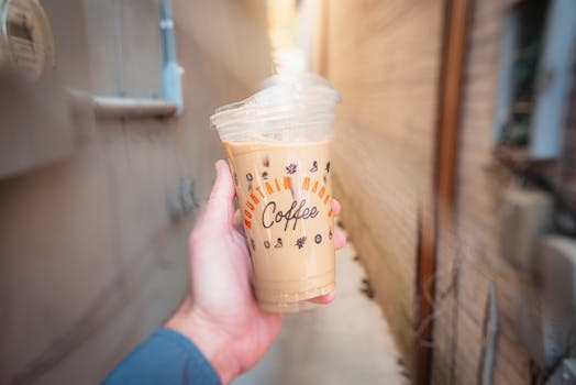 A hand holds an iced coffee with decorative cup branding in a narrow urban alleyway.