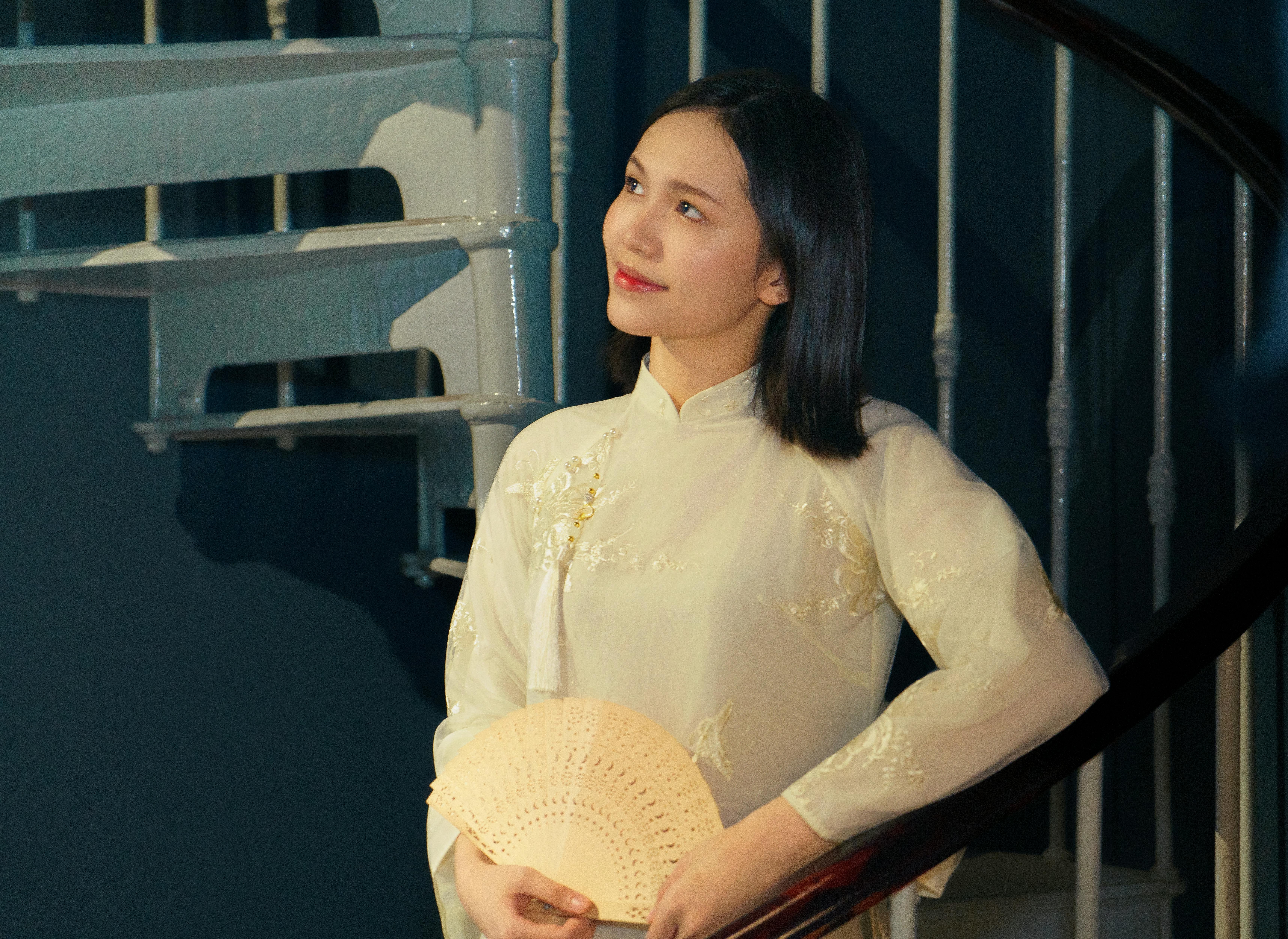 Portrait of a Vietnamese woman in ao dai, holding a fan by a spiral staircase with moody lighting.