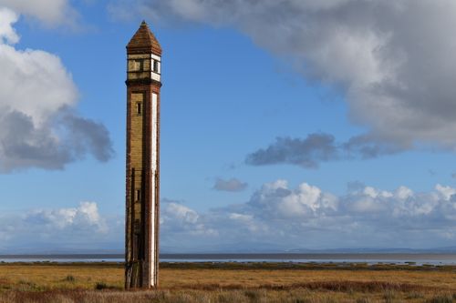 Free Historic brick tower stands solitary in a scenic coastal meadow against a backdrop of a cloudy blue sky. Stock Photo