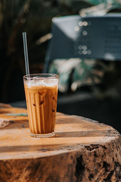 Free A tall glass of iced coffee with straw placed on a rustic wooden table outdoors. Stock Photo
