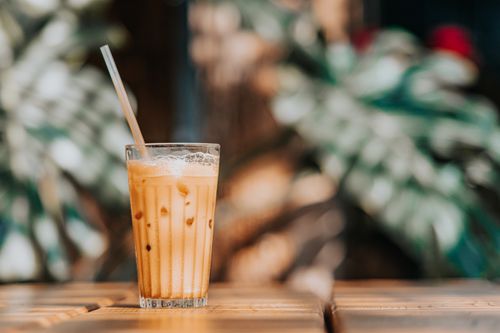 Free Delicious iced coffee in a glass with straw on wooden table against blurred plant background. Stock Photo