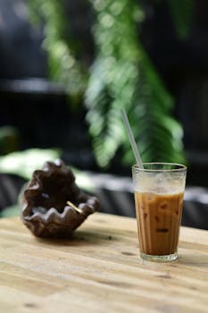Glass of iced coffee with straw on a wooden table, surrounded by lush greenery.