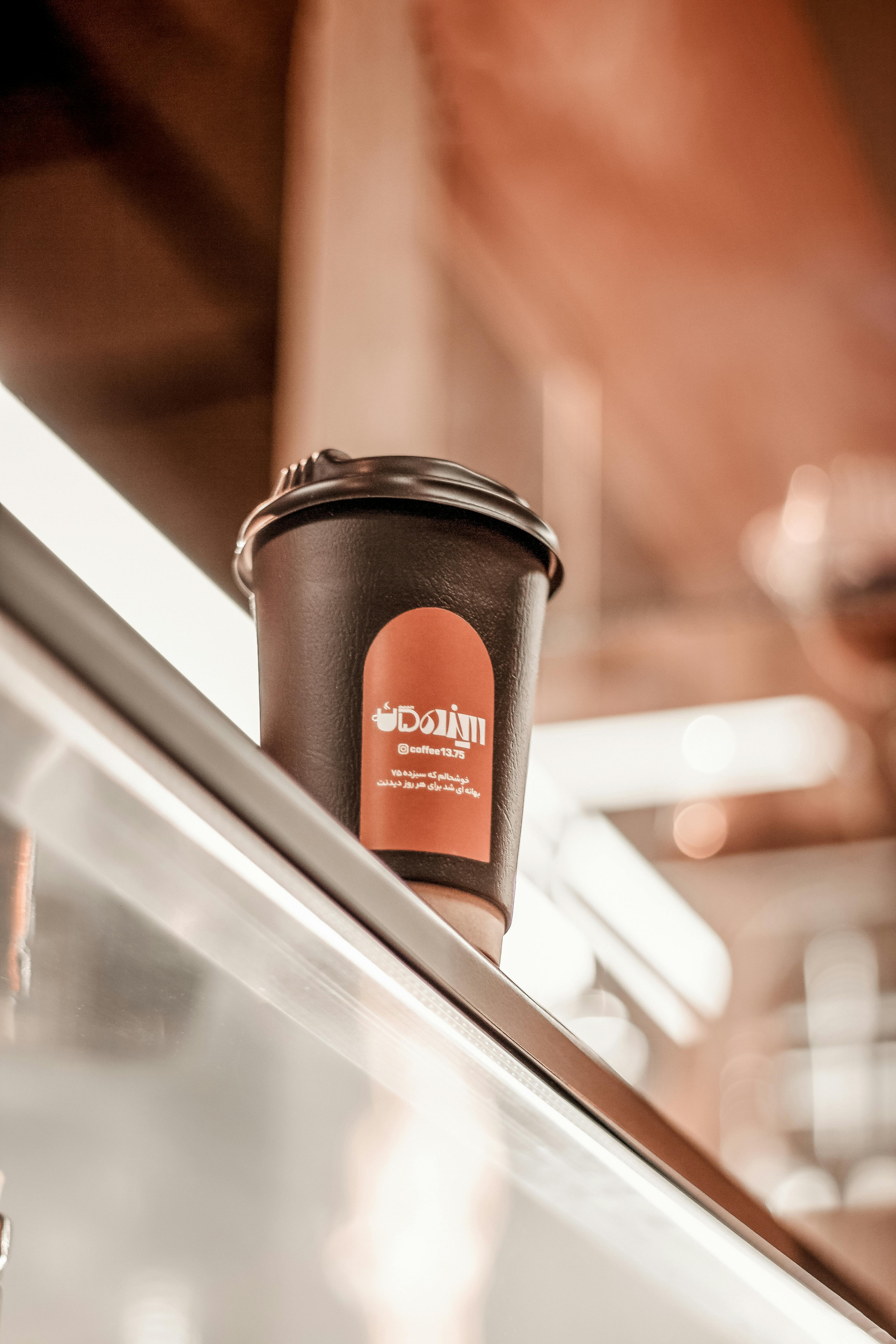 A black takeaway coffee cup with a red logo sits on a cafe shelf in Kermanshah, Iran, creating a cozy atmosphere.