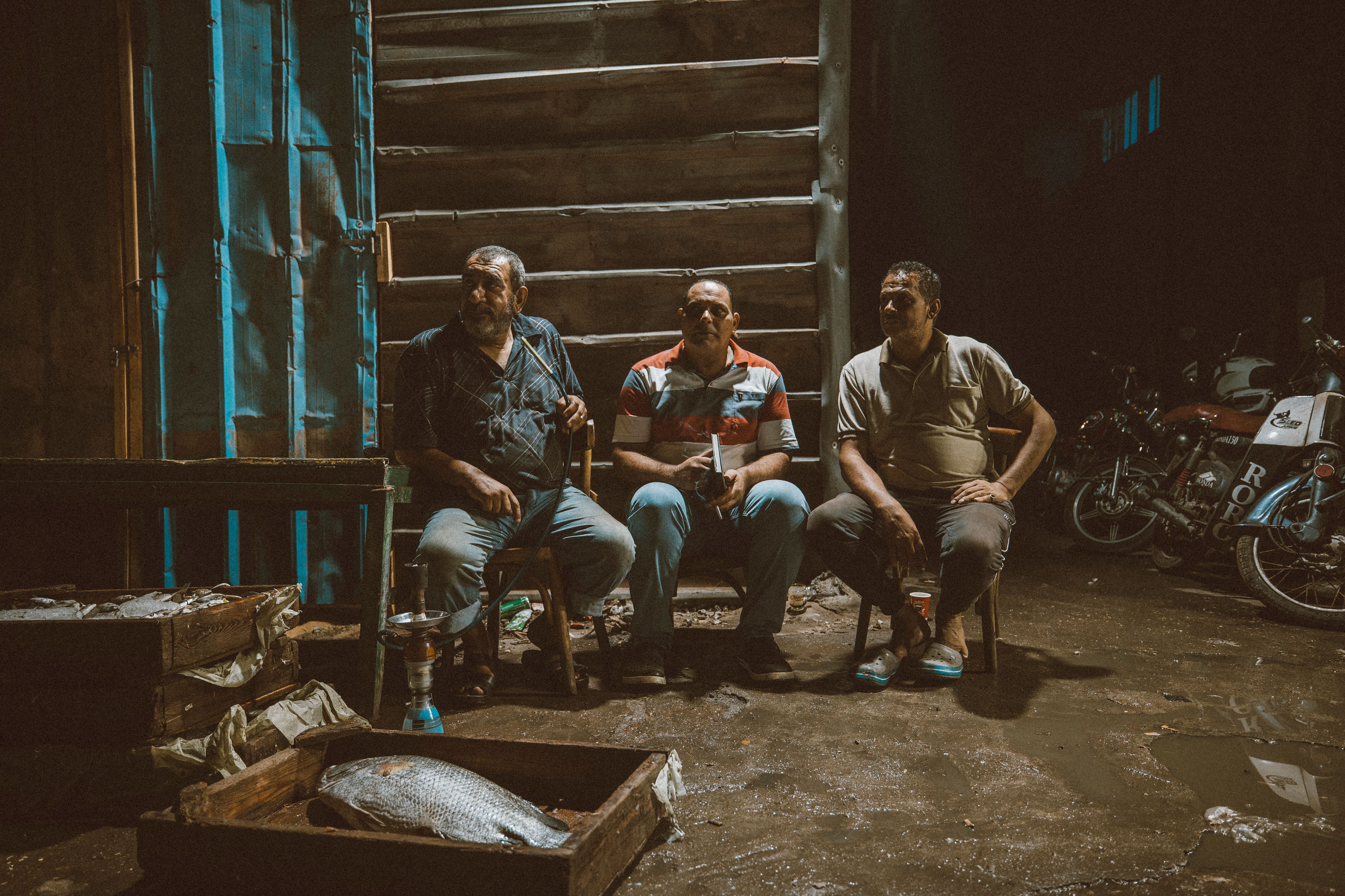 Three men sitting at a fish market in Ras El-Bar, Egypt, with motorcycles in the background.