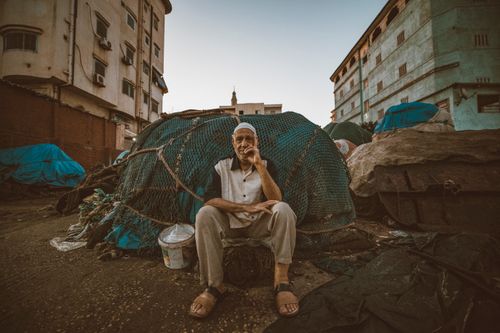 Free An elderly fisherman sits surrounded by fishing nets in Ras El-Bar, Egypt, at dusk, showcasing a scene of local resilience. Stock Photo